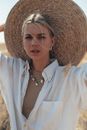 Serious female with tucked back hair wearing white shirt looking at camera while standing with straw hat behind head in nature