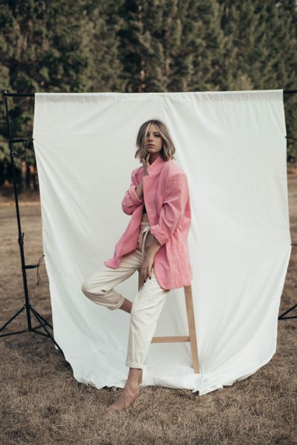 Full body of stylish female model in pink jacket sitting on wooden stool on white background in nature with green trees
