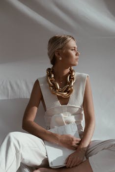 A stylish woman in a white outfit and large gold necklace looks contemplatively to the side.