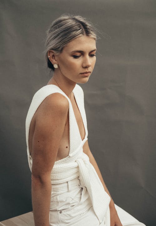 Thoughtful female with tucked back hair in white clothes wearing earrings looking down while sitting on chair on gray background