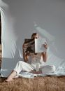 Full body of barefoot female with bent leg sitting in nature on white background and covering half face with magazine