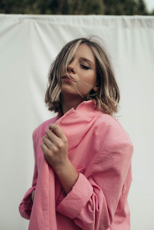 Dreamy female with short hair and closed eyes with hand on pink jacket standing near white background in nature outside