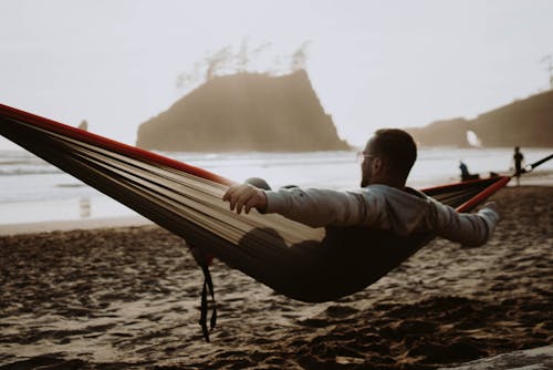 Back view of anonymous male sitting in hammock on sandy beach near sea with rocky cliff in evening time while admiring picturesque view