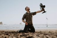 Man on knees on sandy shore near rippling water with table fan in hand while cooling in hot weather in nature
