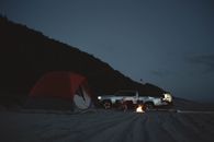 Anonymous tourists on sandy beach with pick up and burning bonfire near camping tent in night time in nature during campaign