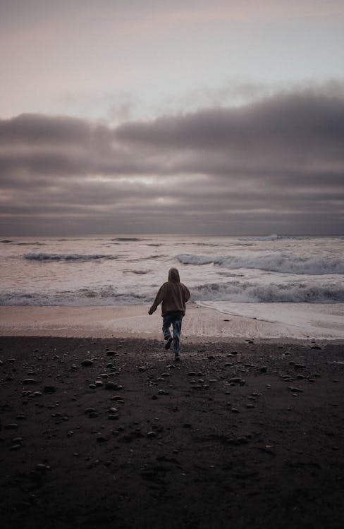 Back view of anonymous person in warm clothes running towards waving sea against cloudy sky in evening