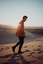 Full length of trendy man walking on sand in desert and looking at camera in sunset back lit