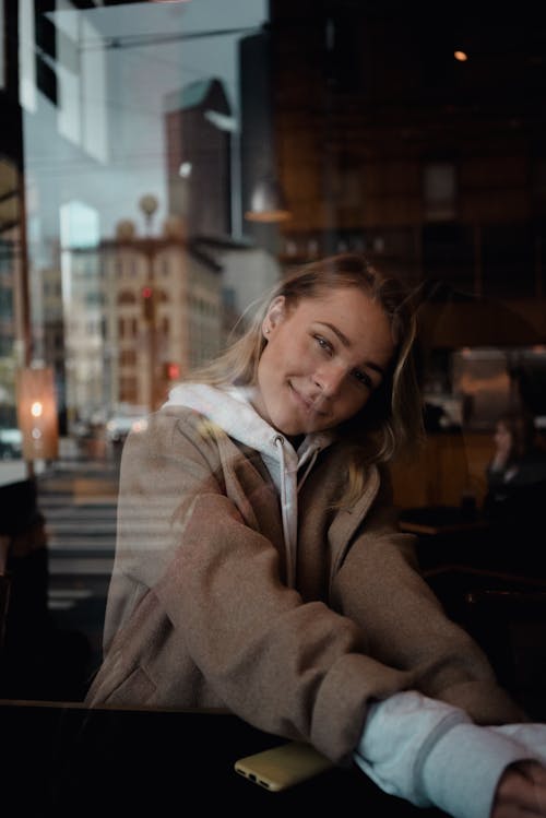 Through window of content blond female in hoodie smiling at camera in coffee shop