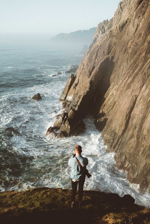 From above of anonymous male admiring powerful water of ocean while standing on hill covered with moss near rough cliff