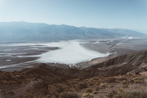 Lake with streams running through dry land with shrubs and rough peaks under hazy sunny sky