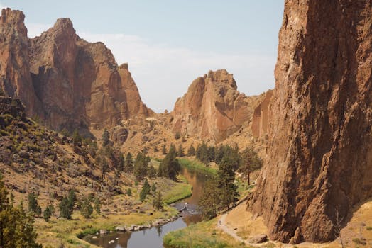 Narrow lake flowing among grassy terrain with trees surrounded with rocky cliffs against cloudy sky in national park on summer day