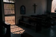 Dark chapel with cross on wall and wooden benches located in city with metal gate casting shadow on floor on sunny day
