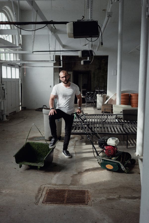 Full body of male worker looking at camera while standing in garage near lawn mower during preparation for cutting grass