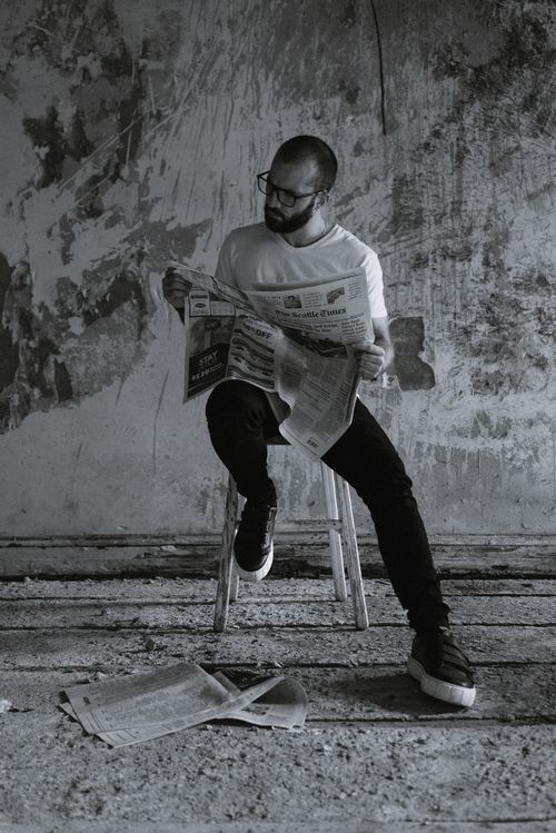 Free Black and white full body of focused bearded male in eyeglasses reading newspaper while sitting on chair against shabby wall in neglected building Stock Photo