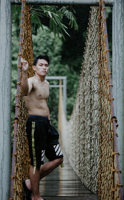 Young attentive ethnic male in shorts with naked torso standing on aged bridge while looking at camera