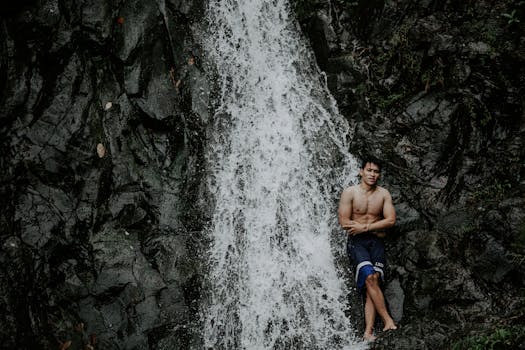 Young fit shirtless ethnic male traveler standing with crossed legs and arms near rapid cascade in mounts