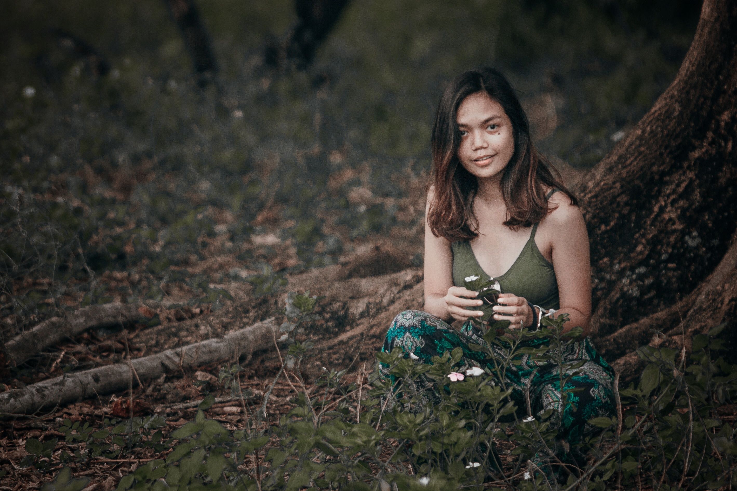 Young content ethnic female sitting with crossed legs near tree with blossoming flowers and looking at camera