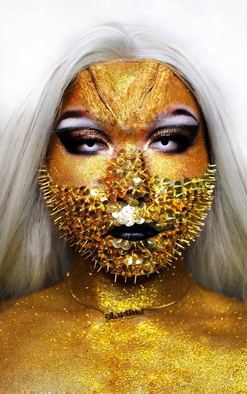 Woman with white hair covered in shiny golden paint looking at camera while standing on white background in mask with spikes