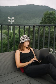 A woman in a black tank top and leopard-print hat works on her laptop outdoors, enjoying remote work freedom.