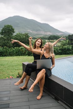 Two women enjoying outdoor leisure by the pool with a laptop, set against a scenic mountain backdrop.