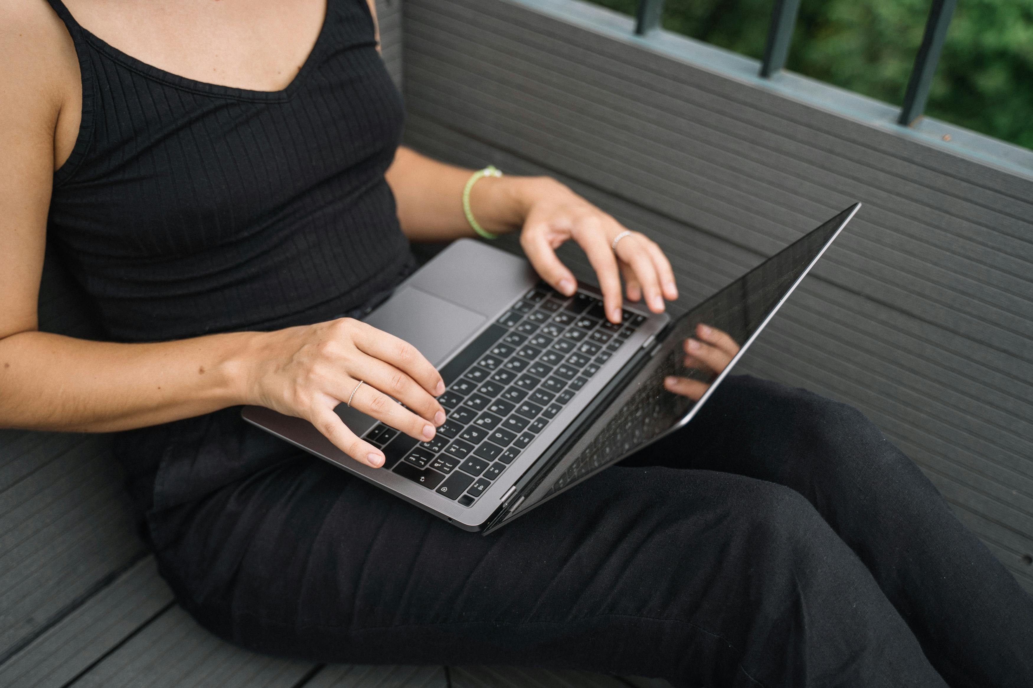 Close-up of woman typing on laptop while sitting outdoors on a wooden deck.