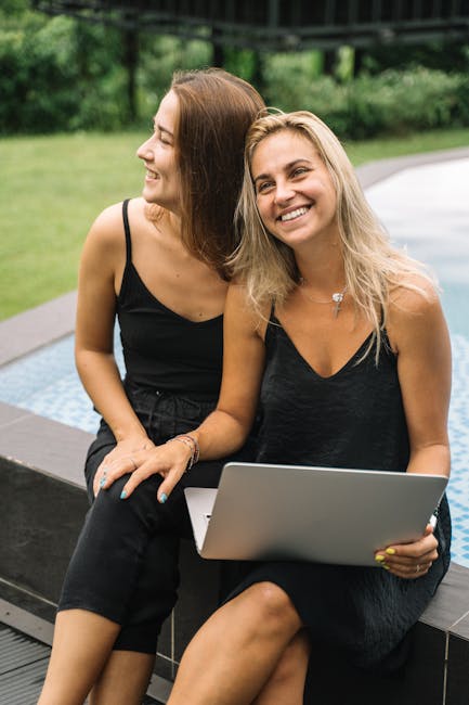 Two smiling women relaxing by a pool with a laptop, enjoying a sunny day outdoors.