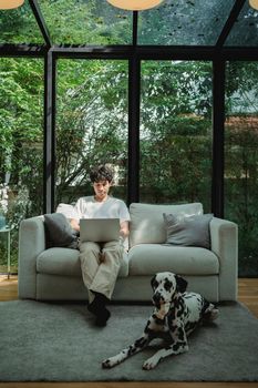 Man sitting on couch using laptop with Dalmatian dog nearby. Cozy indoors setting with greenery outside.