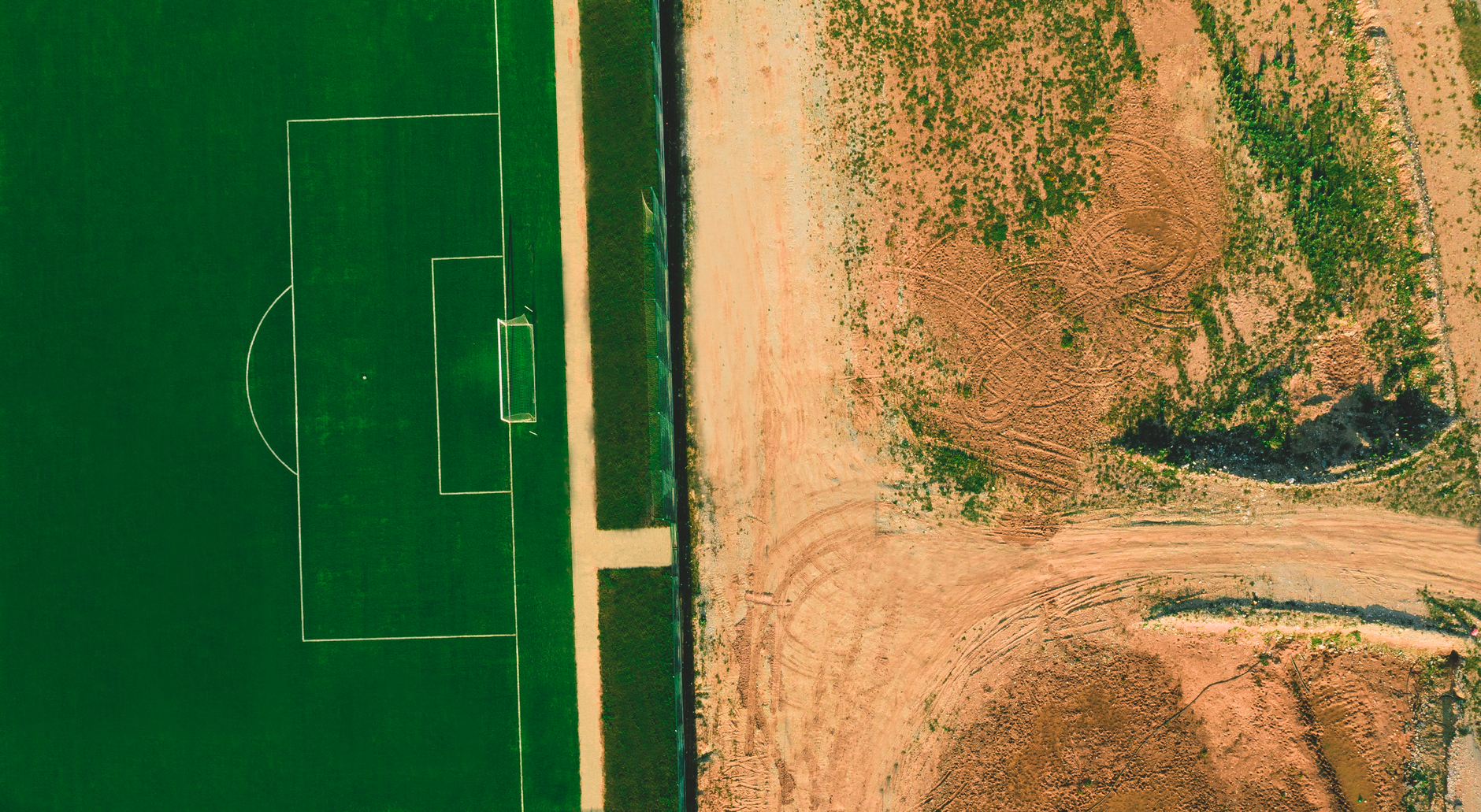Top-down aerial view of a green football pitch next to a barren dirt field.