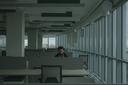 一名成年男子独自坐在开放式办公室里，四周是办公桌和隔板