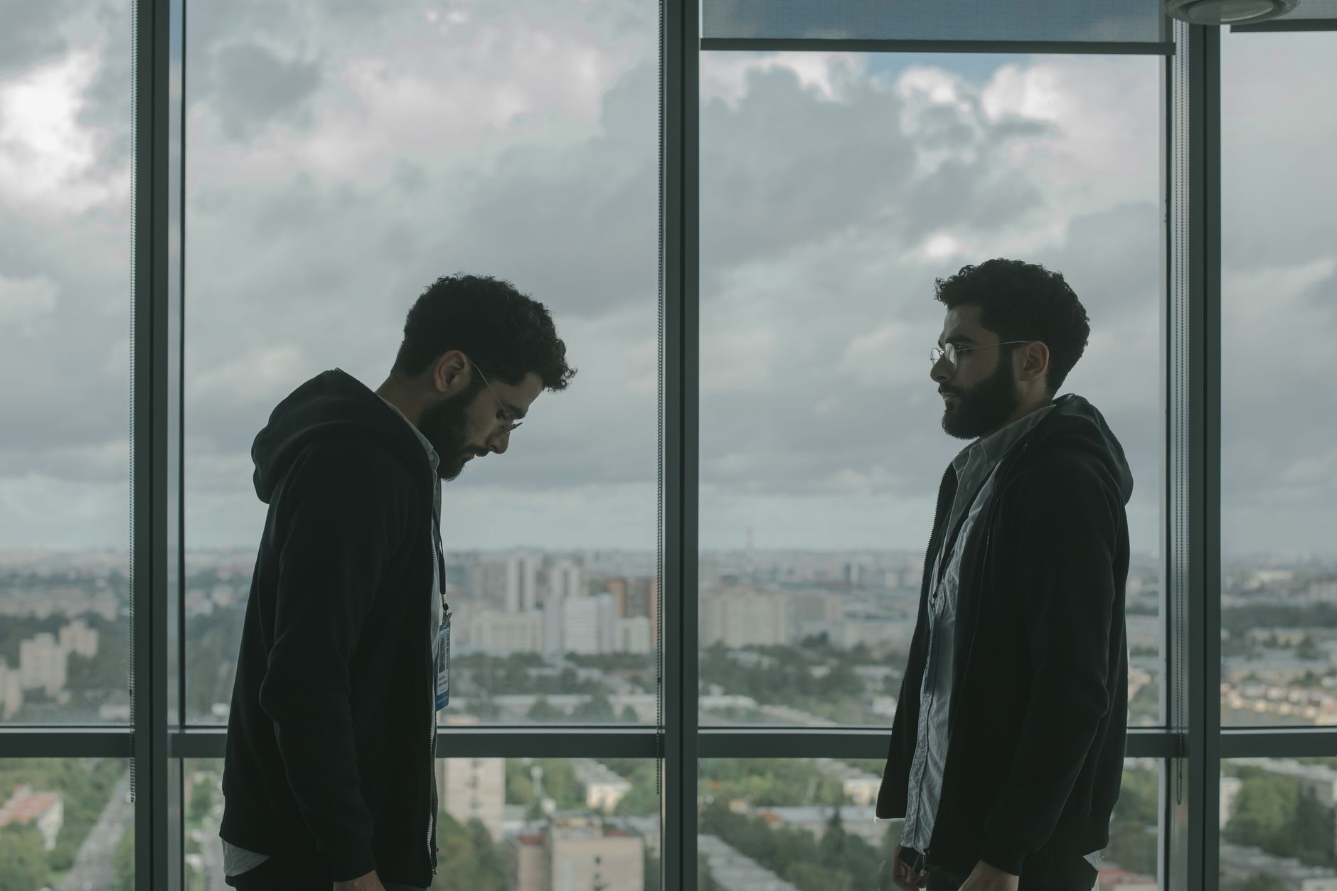 Two men stand facing each other by a window overlooking a cityscape. Moody ambiance.