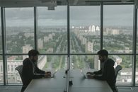 在现代办公室里，两名男子正对着笔记本电脑工作，透过大窗户可以看见城市的全景
