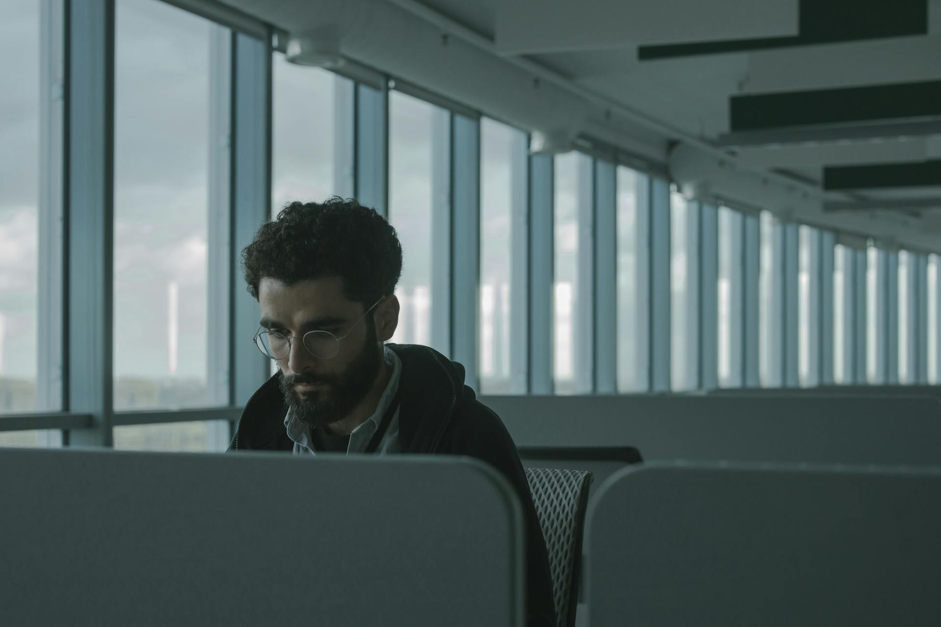 A man with glasses working in a spacious modern airport lounge with large windows.