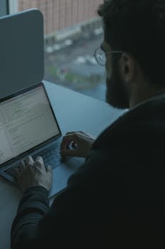 A man coding on his laptop by a window in an office setting, showcasing technology work.