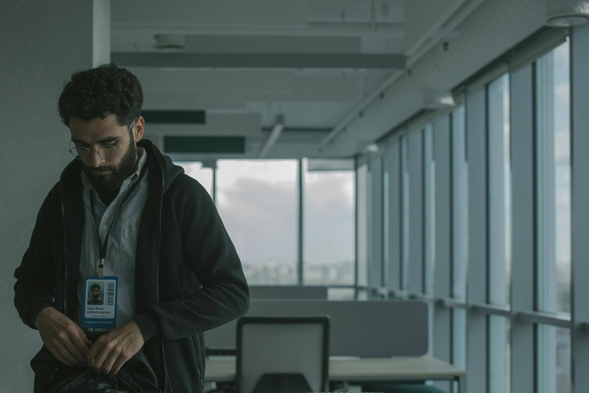 Man wearing eyeglasses and ID badge in modern office setting. Office interior with large windows.