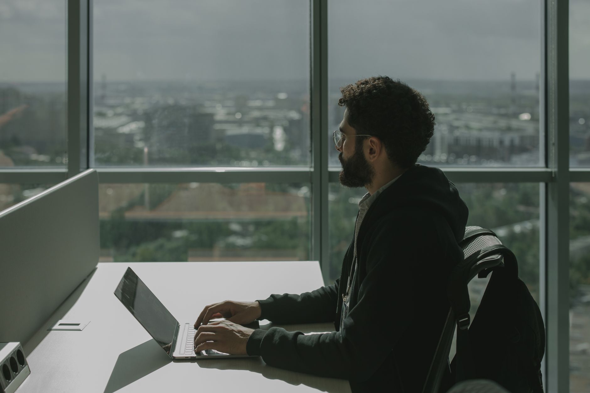 A man with facial hair types on a laptop at a modern office desk with a city view.
