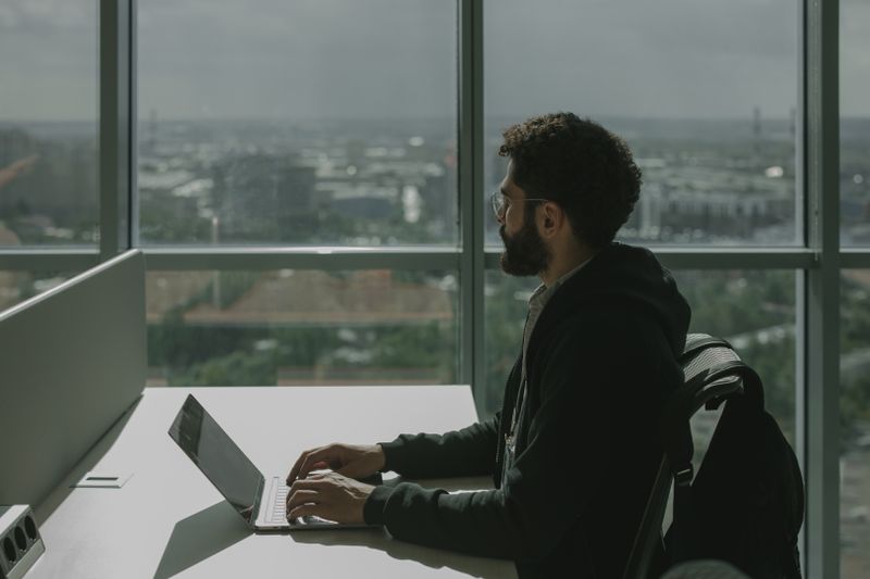 A man with facial hair types on a laptop at a modern office desk with a city view.