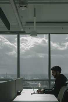 Man in a black jacket sitting at a desk and working on a laptop in a bright modern urban office with glass windows.