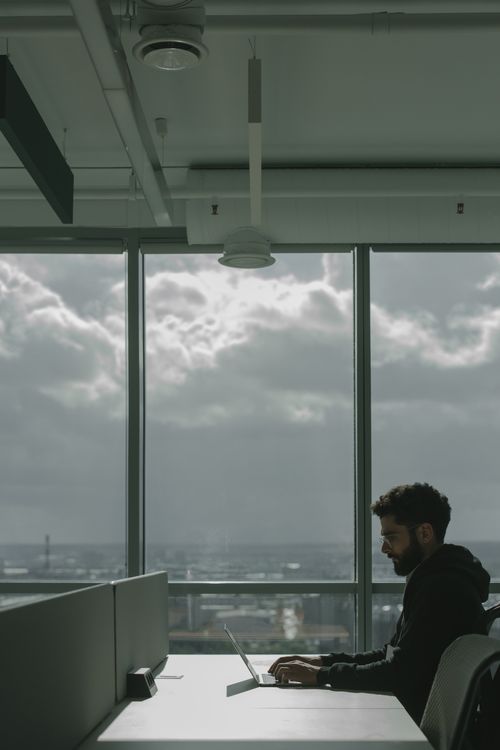 Free Man in a black jacket sitting at a desk and working on a laptop in a bright modern urban office with glass windows. Stock Photo