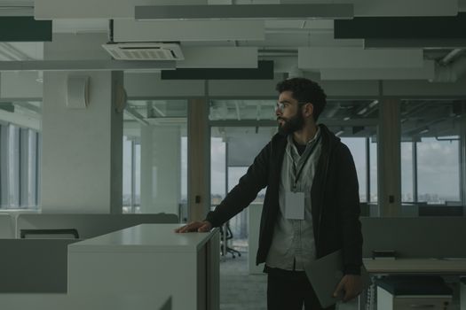 Business professional in a modern office with smart attire and a laptop, gazing thoughtfully.