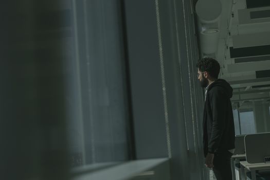 一个身着连帽衫的男人站在办公室里，侧身凝视着窗外
