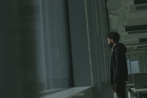 Free Side view of a man in a hoodie standing in an office, gazing out the window. Stock Photo