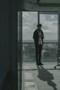 一名身穿黑色夹克的男子站在办公室的大窗户旁，俯瞰着城市的天际线