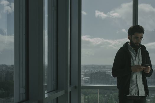 一名男子站在现代办公室的玻璃窗旁，用智能手机映出窗外的城市景观