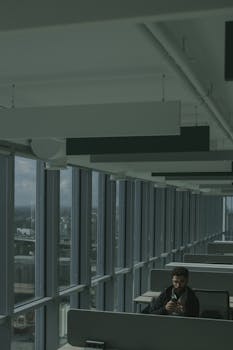 A man sits alone using his phone in a modern office with expansive windows.