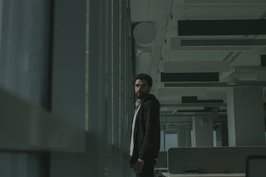 A bearded man in a dark jacket stands thoughtfully by a window in a modern office environment.