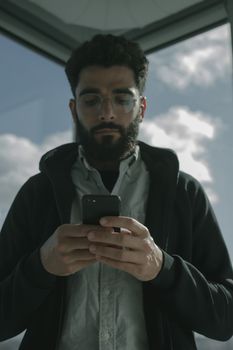 Bearded man in eyeglasses using smartphone indoors, focused on device.