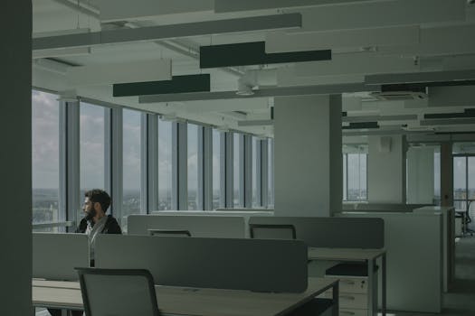 A man sits alone in a modern office space with large windows overlooking a cityscape.