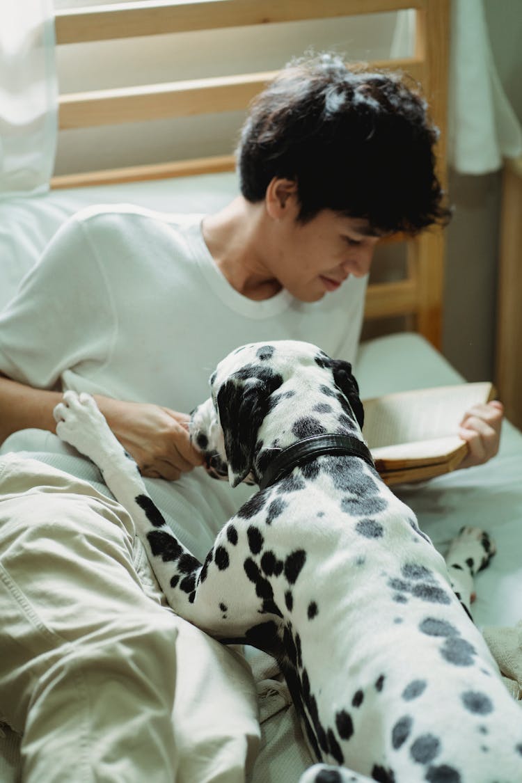 A Man Reading A Book On His Bed With His Dog