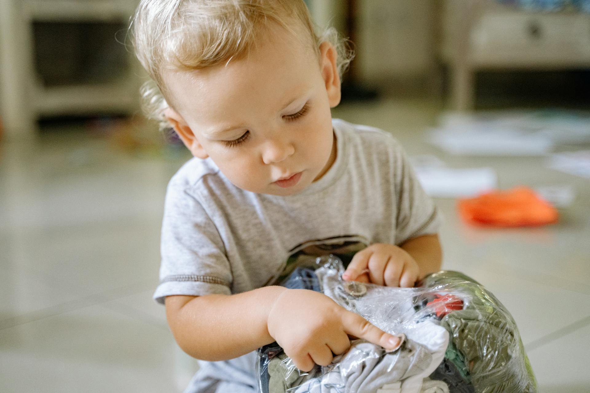 A toddler playing with clothes wrapped in a plastic bag indoors, showcasing curiosity and focus.
