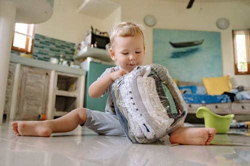 Free Curious toddler sitting on the floor playing with a plastic bag of clothes indoors. Stock Photo
